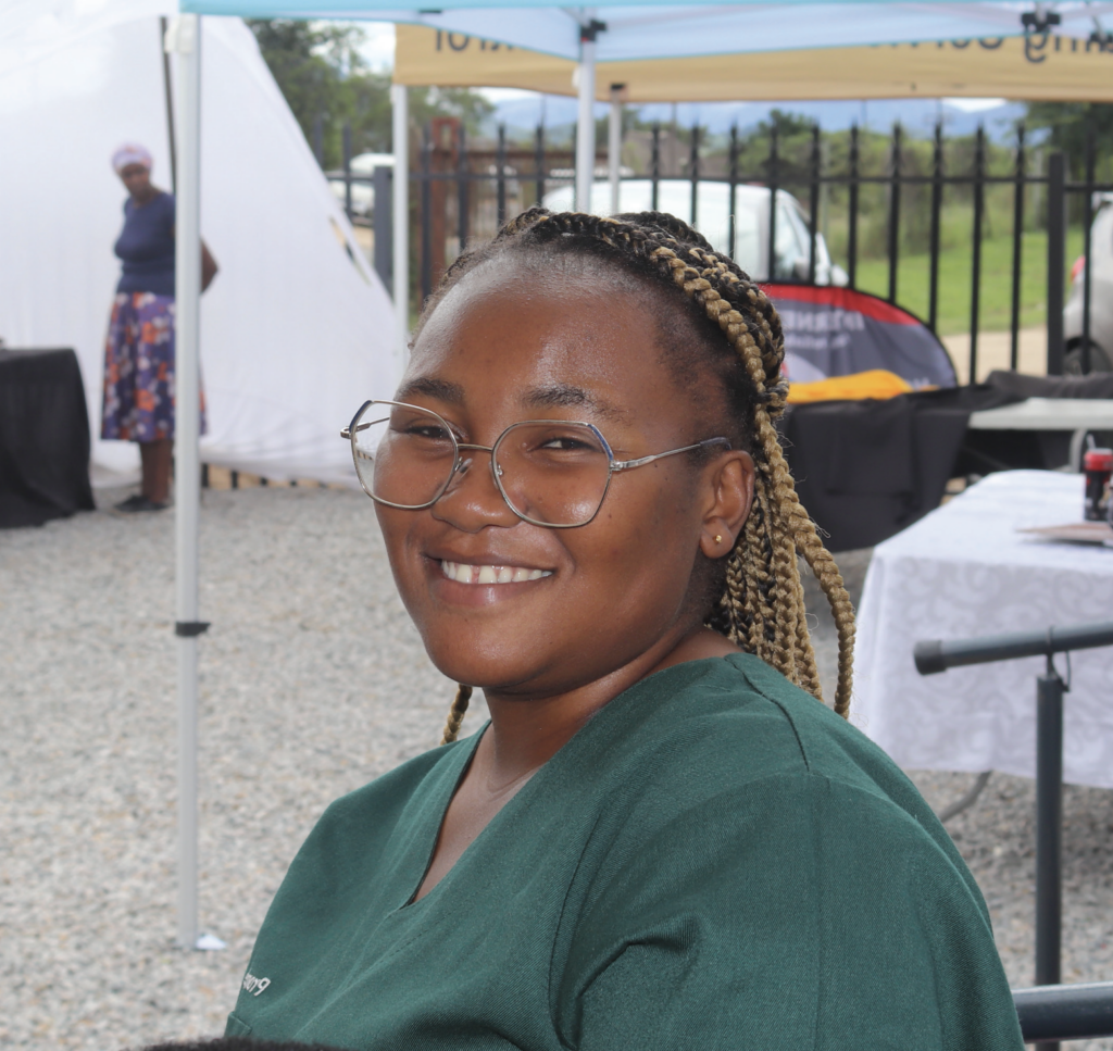 African woman sitting under medical tent, smiling at camera and wearing green scrub top and glasses.